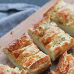 Close-up of sliced baked bread with garlic and cheese topping, placed on a wooden cutting board, with a folded napkin in the background.