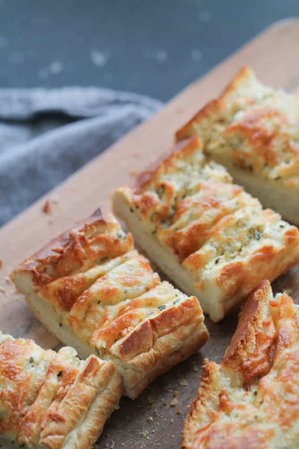 Close-up of sliced baked bread with garlic and cheese topping, placed on a wooden cutting board, with a folded napkin in the background.