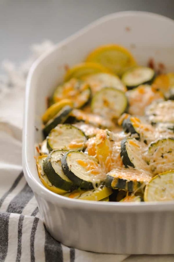 A baked dish of sliced zucchini and yellow squash topped with melted cheese in a white casserole dish, placed on a striped cloth.