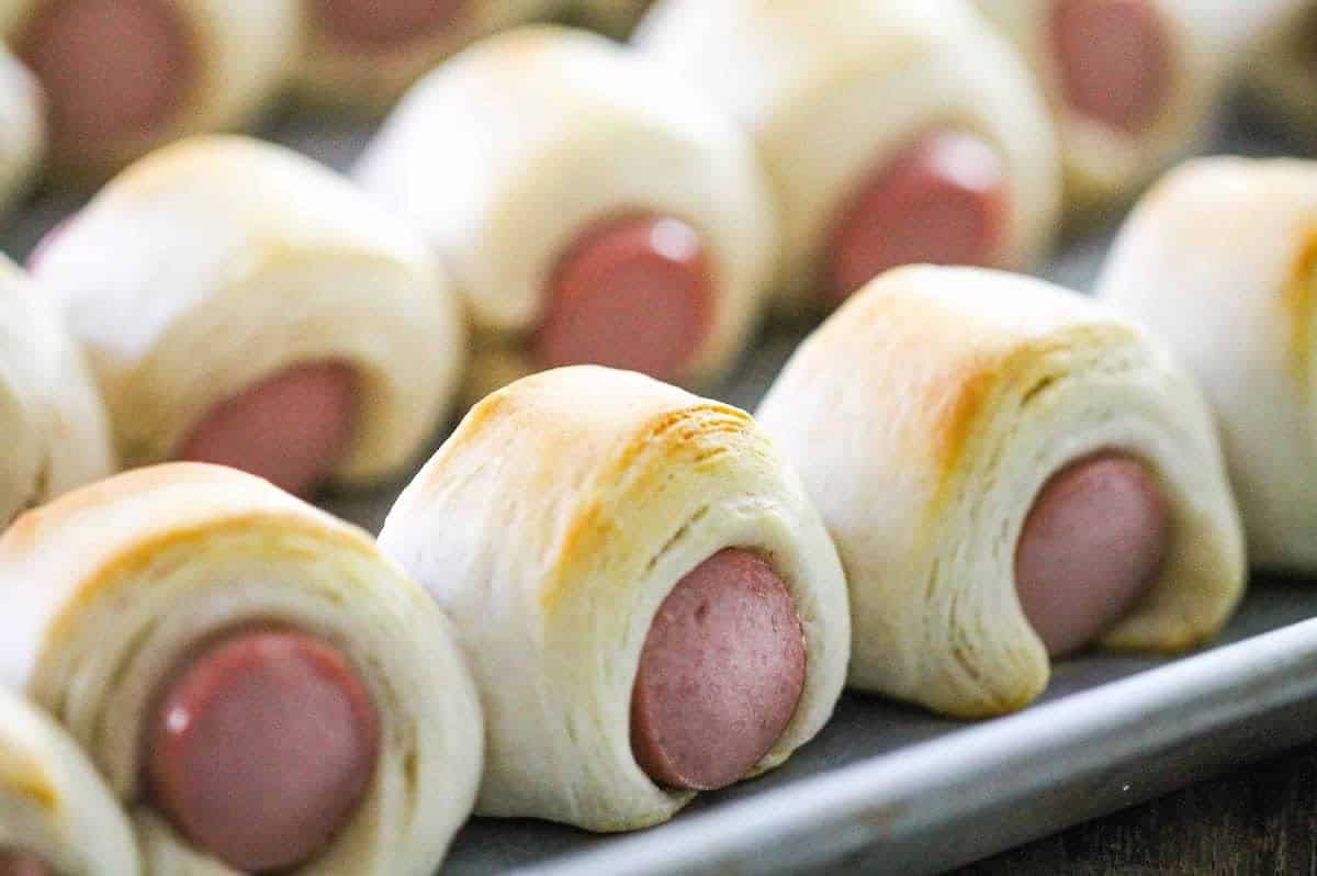 Close-up of several pigs in a blanket appetizers arranged on a baking sheet. The golden-brown dough wraps around small hot dog sausages, with some edges slightly browned from baking.