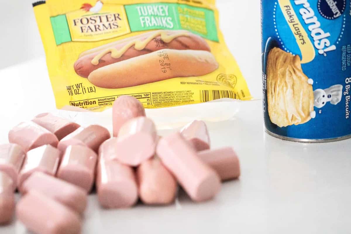 Canned biscuits, a package of turkey franks, and several sliced hot dogs on a white surface.