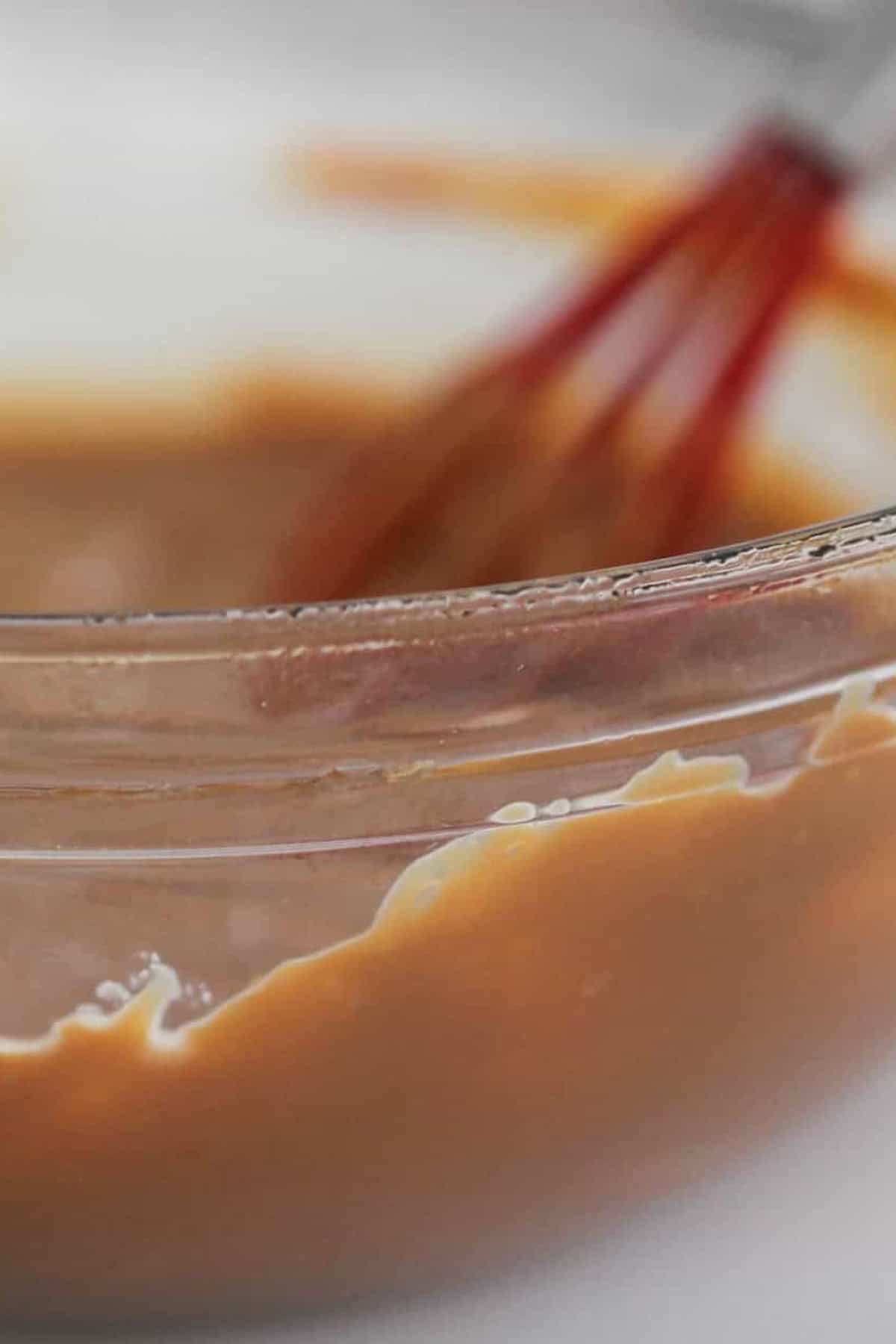 A close-up of a glass bowl filled with light brown batter, with a red whisk partially visible in the background.