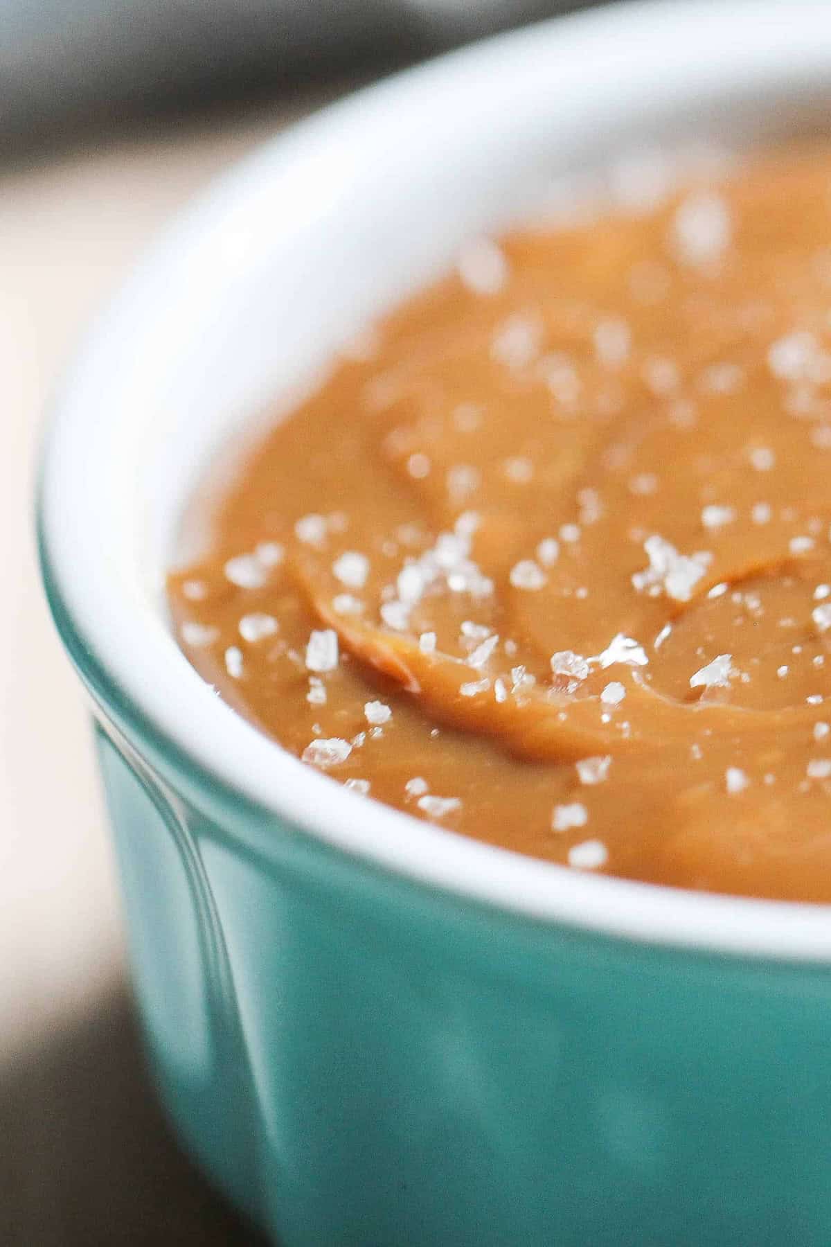 Close-up of salted caramel sauce in a light blue ceramic dish, topped with coarse sea salt crystals.