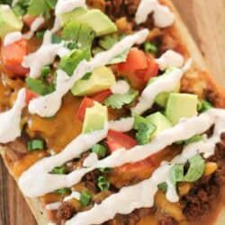 Rectangular flatbread topped with ground meat, melted cheese, diced tomatoes, avocado, cilantro, green onions, and drizzled with white sauce.