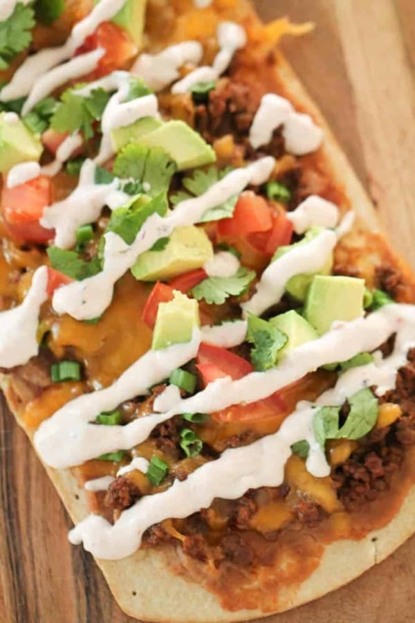 Rectangular flatbread topped with ground meat, melted cheese, diced tomatoes, avocado, cilantro, green onions, and drizzled with white sauce.