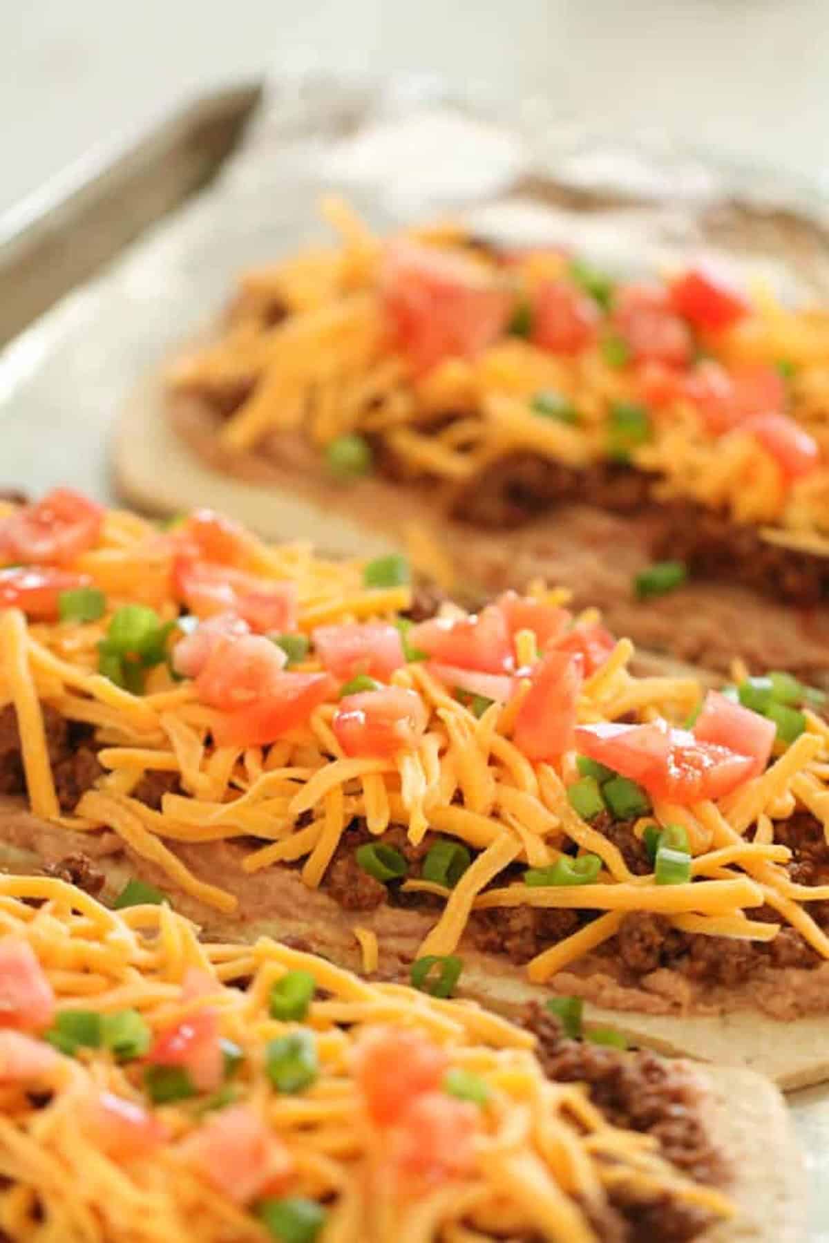 Flatbreads topped with ground meat, shredded cheddar cheese, diced tomatoes, and chopped green onions are arranged on a tray.