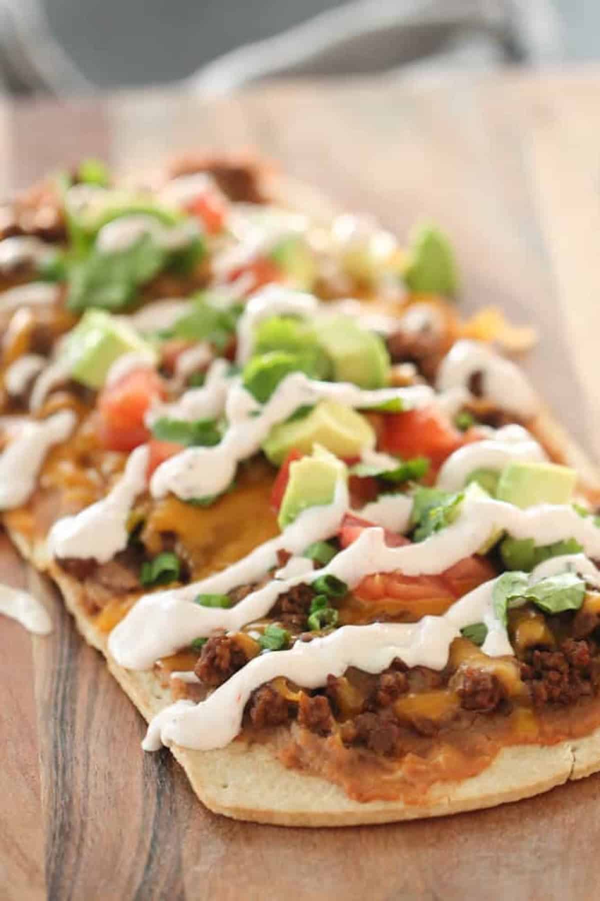 Rectangular flatbread topped with ground meat, melted cheese, diced tomatoes, avocado, green onions, and drizzled with white sauce on a wooden surface.