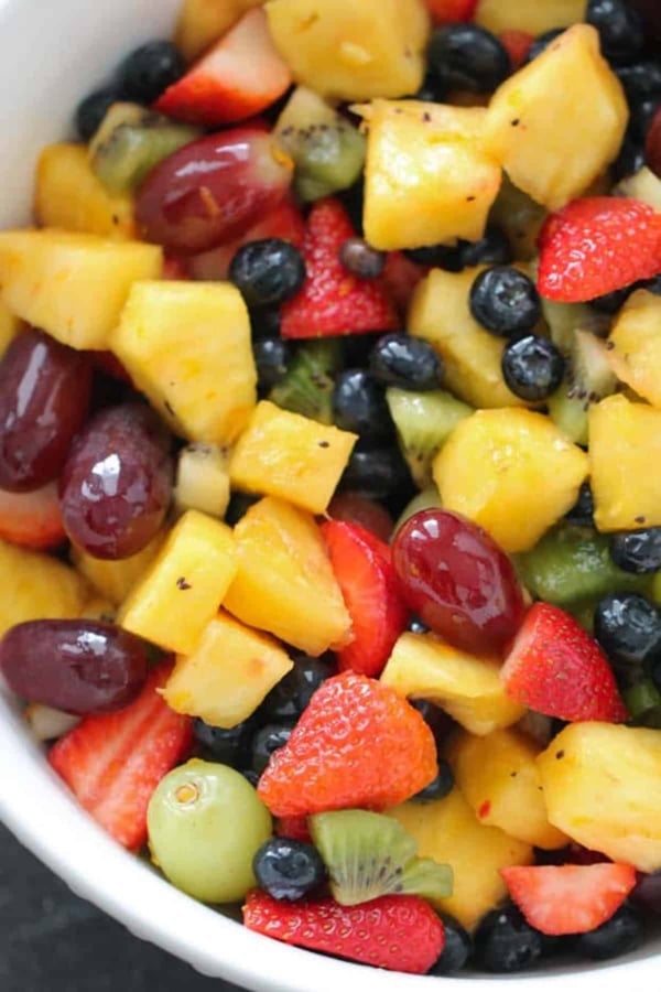 A bowl filled with a mixed fruit salad containing pineapple chunks, strawberries, blueberries, grapes, and kiwi slices.