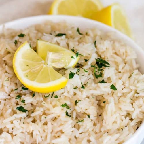 A white bowl filled with cooked rice, garnished with chopped herbs and two lemon wedges on top, with two lemon halves in the background.