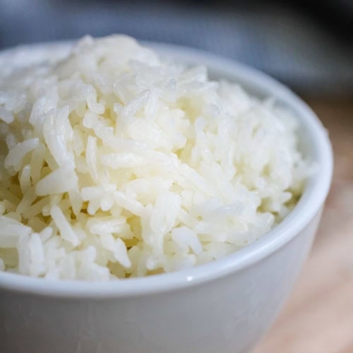 A white ceramic bowl filled with cooked white rice sits on a wooden surface.