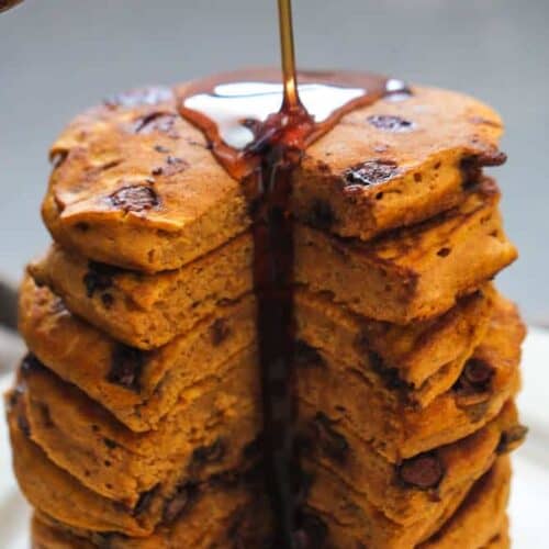 A stack of thick pancakes with visible chocolate chips being drizzled with syrup.