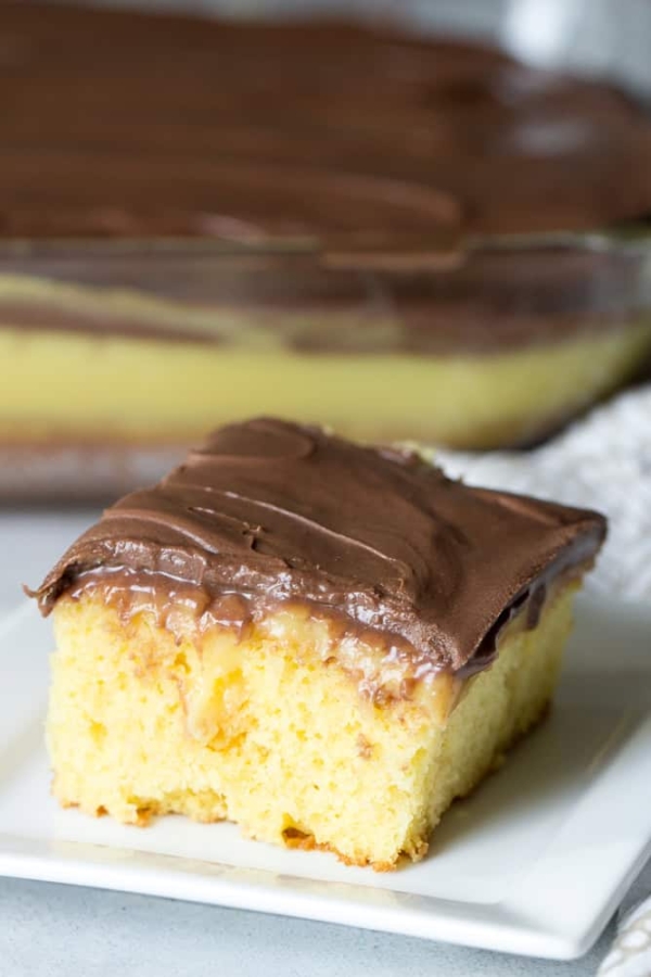 A slice of Boston Cream Poke Cake on a white plate with the remaining cake in a pan in the background.