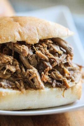 A close-up of a pulled pork sandwich served on a white plate. The sandwich has shredded pork meat inside a bun.