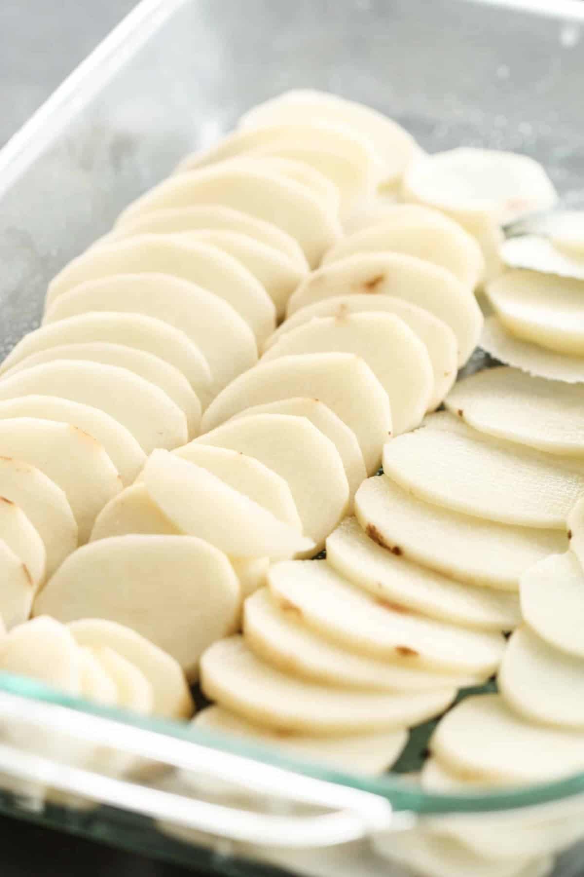 Thinly sliced raw potatoes are arranged in overlapping rows in a glass baking dish.