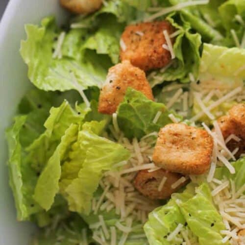 Close-up of a Caesar salad with romaine lettuce, shredded Parmesan cheese, and seasoned croutons in a white bowl.