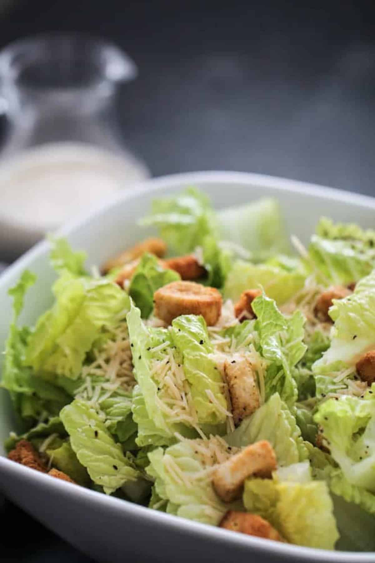 A bowl of Caesar salad with romaine lettuce, croutons, shredded Parmesan cheese, and a glass container of dressing in the background.