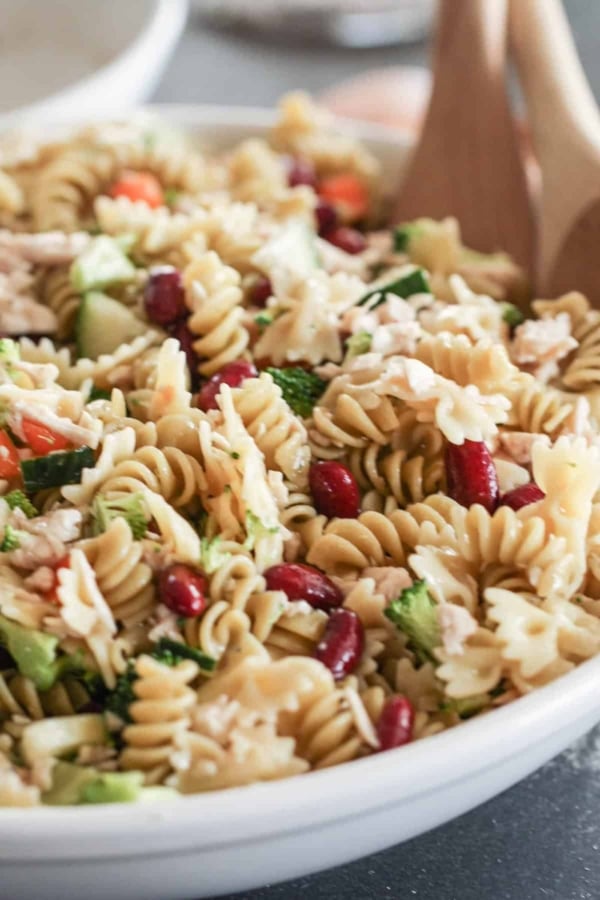 A bowl of pasta salad with rotini, kidney beans, chopped vegetables, and shredded chicken, accompanied by wooden serving utensils.