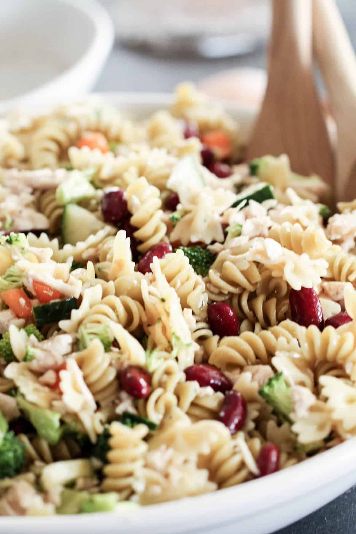 A bowl of rotini pasta salad with red kidney beans, diced vegetables, and wooden serving utensils.