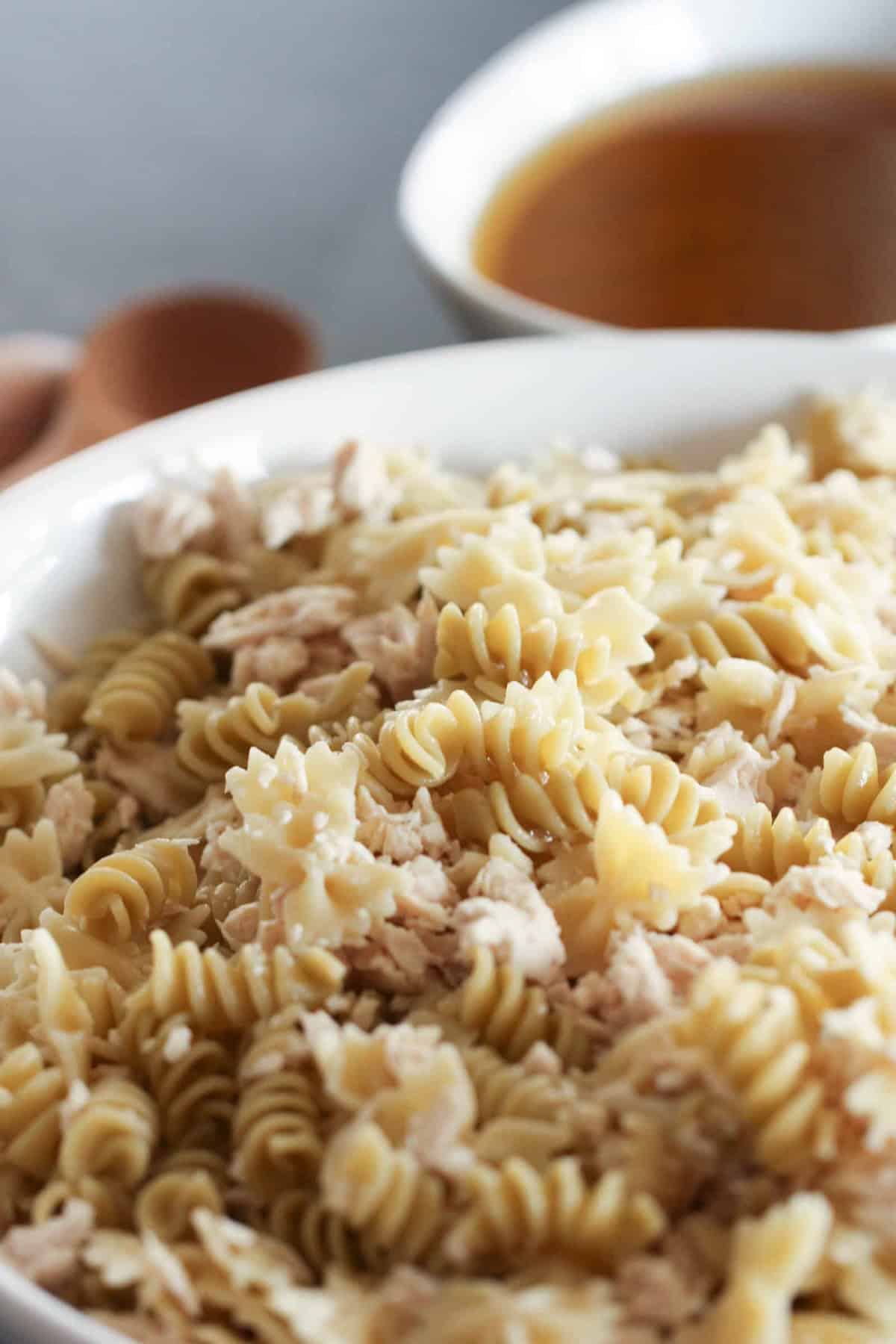 Close-up of a bowl containing rotini pasta mixed with shredded chicken, with a bowl of broth partially visible in the background.