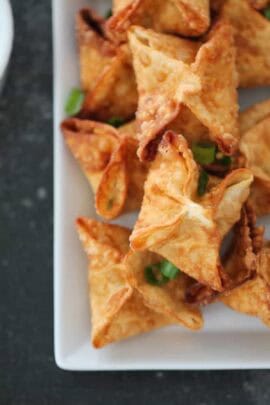 A plate of golden-brown wontons garnished with chopped green onions on a dark background.