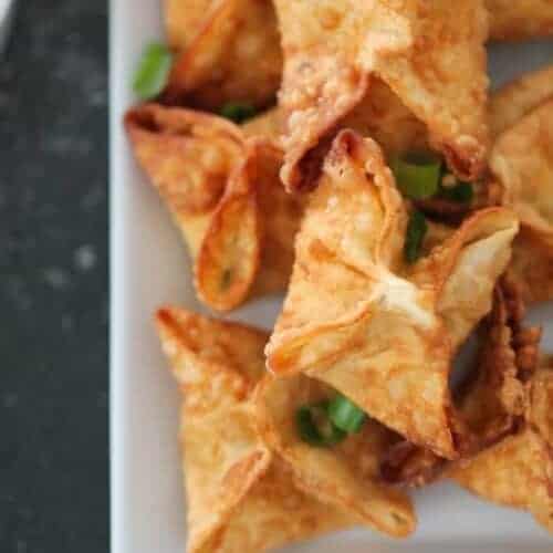 A plate of golden-brown wontons garnished with chopped green onions on a dark background.