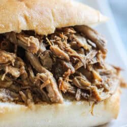 A close-up of a pulled pork sandwich served on a white plate. The sandwich has shredded pork meat inside a bun.
