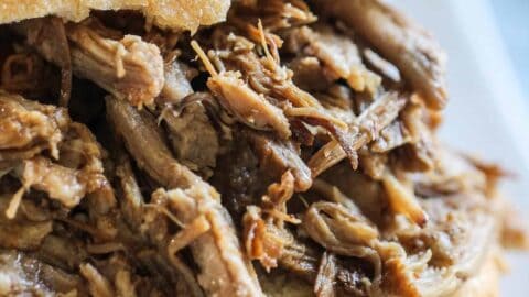 A close-up of a pulled pork sandwich served on a white plate. The sandwich has shredded pork meat inside a bun.
