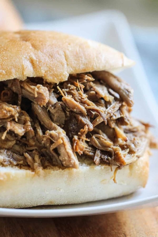 A close-up of a pulled pork sandwich served on a white plate. The sandwich has shredded pork meat inside a bun.