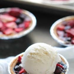 A small fruit tart in a foil tin topped with a scoop of vanilla ice cream, with more similar tarts in the background.