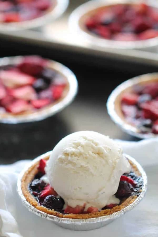 A small fruit tart in a foil tin topped with a scoop of vanilla ice cream, with more similar tarts in the background.