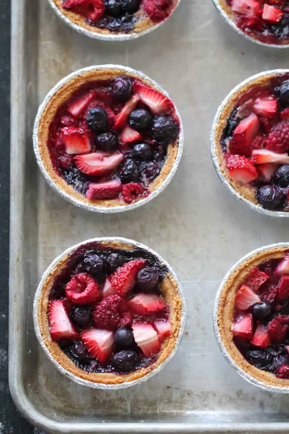 Mini berry pies with strawberries, blueberries, and raspberries in aluminum tins arranged on a metal baking sheet.