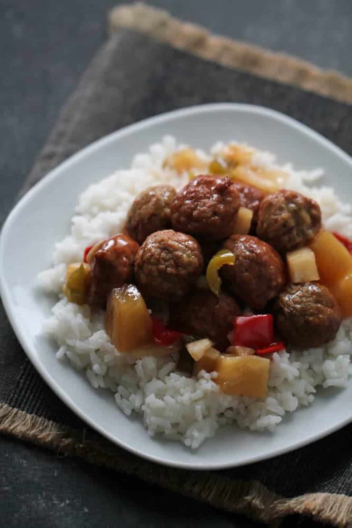 A plate of rice topped with meatballs, pineapple chunks, red and yellow bell peppers in a sauce, served on a white square dish.