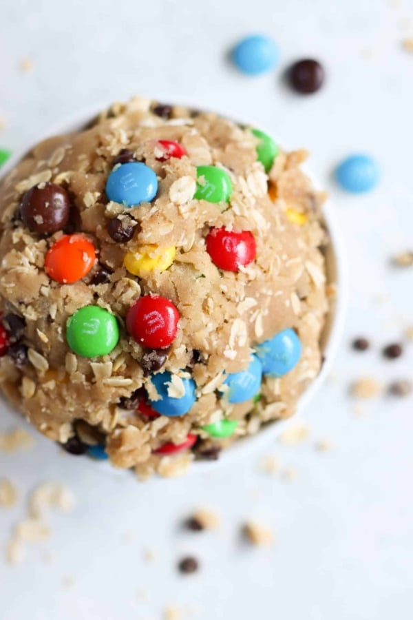 Edible cookie dough mixed with oats, m&ms and chocolate chips. In a small white bowl with m&ms and chocolate chips surround the bowl.