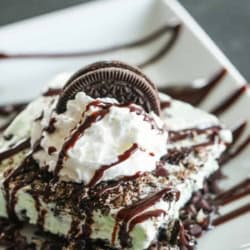 A slice of Asphalt Sheet Cake Pie on a white plate drizzled with chocolate sauce and topped with whip cream and an oreo cookie.