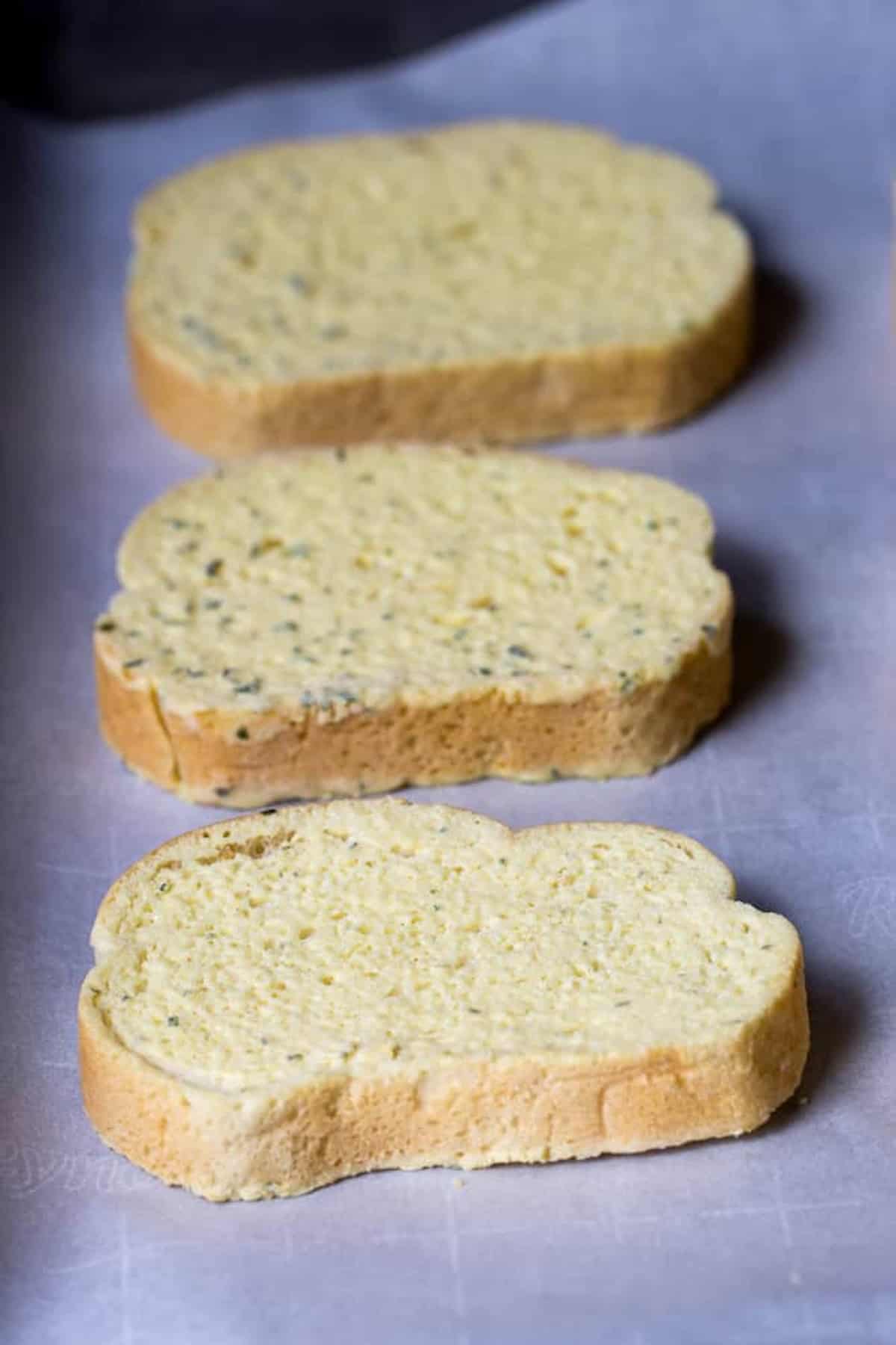 Three slices of plain, uncooked bread are arranged in a row on a sheet of parchment paper.