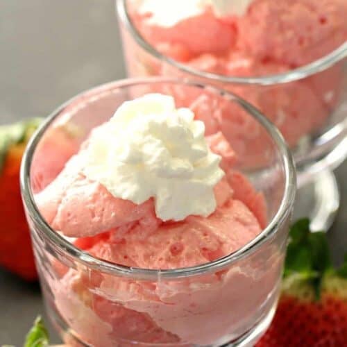 Two glass dishes filled with pink strawberry mousse topped with whipped cream, with fresh strawberries placed beside the dishes.