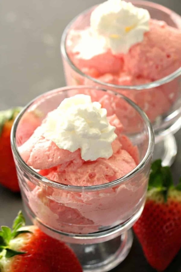 Two glass dishes filled with pink strawberry mousse topped with whipped cream, with fresh strawberries placed beside the dishes.