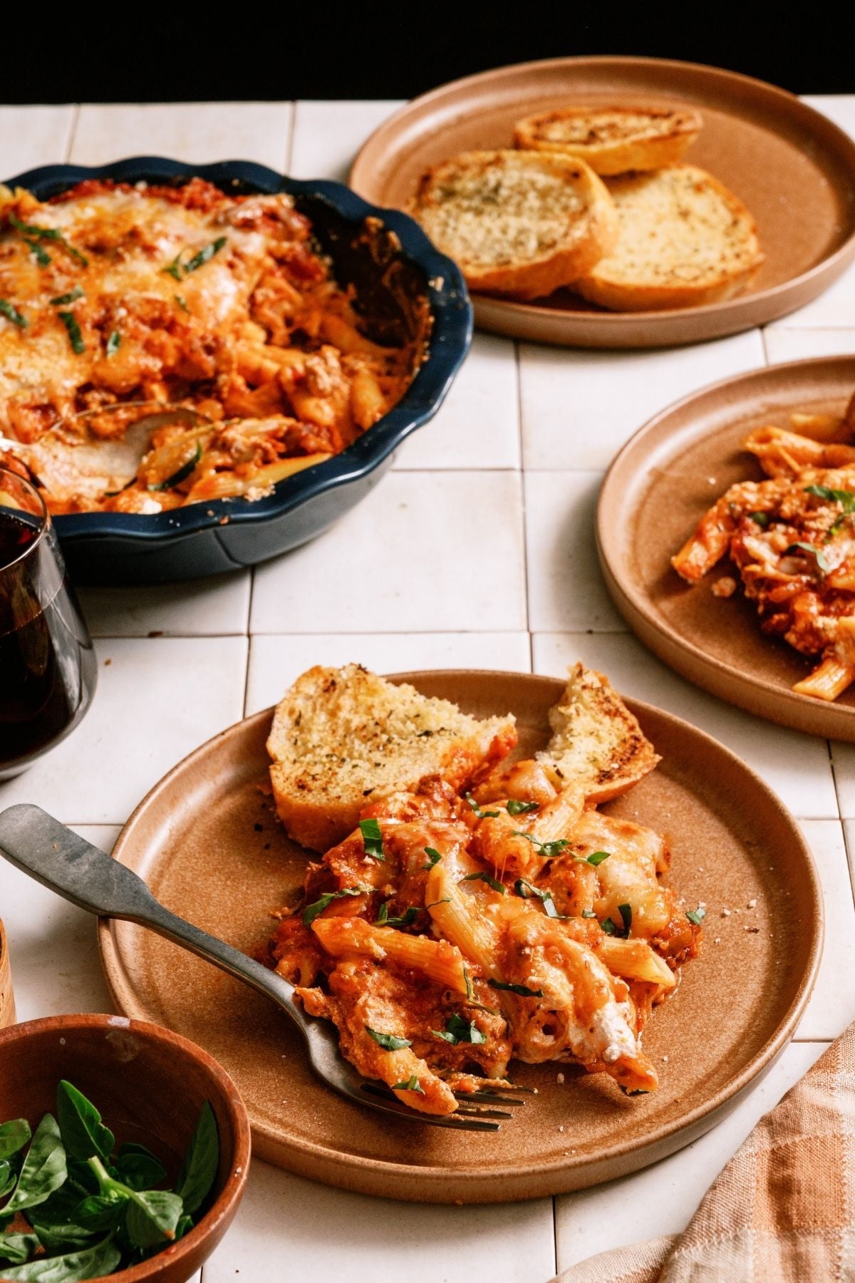 A serving of Baked Ziti on a plate with a slice of garlic bread. The remaining Baked Ziti in the round baking dish.