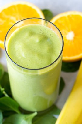 A glass of green smoothie is placed in the center, with orange slices and a banana in the background.