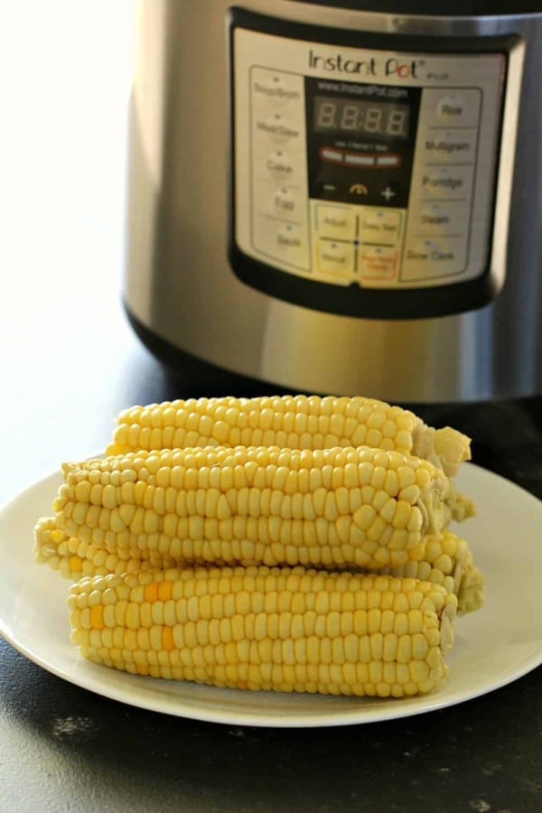 Four ears of corn on a white plate in front of an Instant Pot pressure cooker.