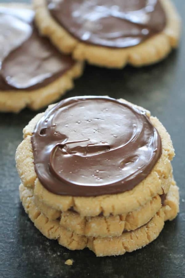 Three peanut butter cookies stacked, each topped with a layer of chocolate frosting, with more cookies in the background.