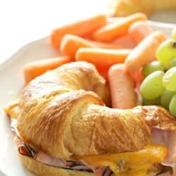 Croissant sandwich with ham and melted cheese on a plate, accompanied by baby carrots and green grapes.