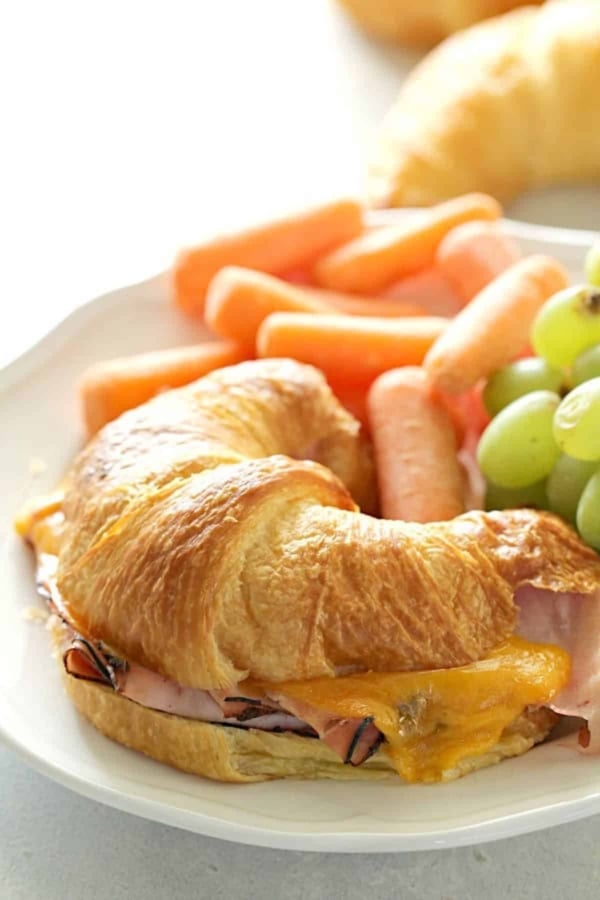 Croissant sandwich with ham and melted cheese on a plate, accompanied by baby carrots and green grapes.