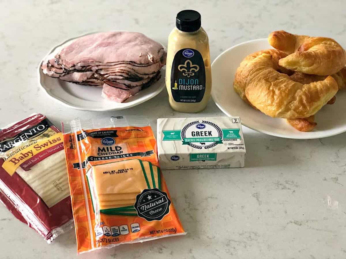 Slices of ham, a plate with two croissants, Dijon mustard, Swiss and mild cheddar cheese packages, and a block of cream cheese are arranged on a marble countertop.