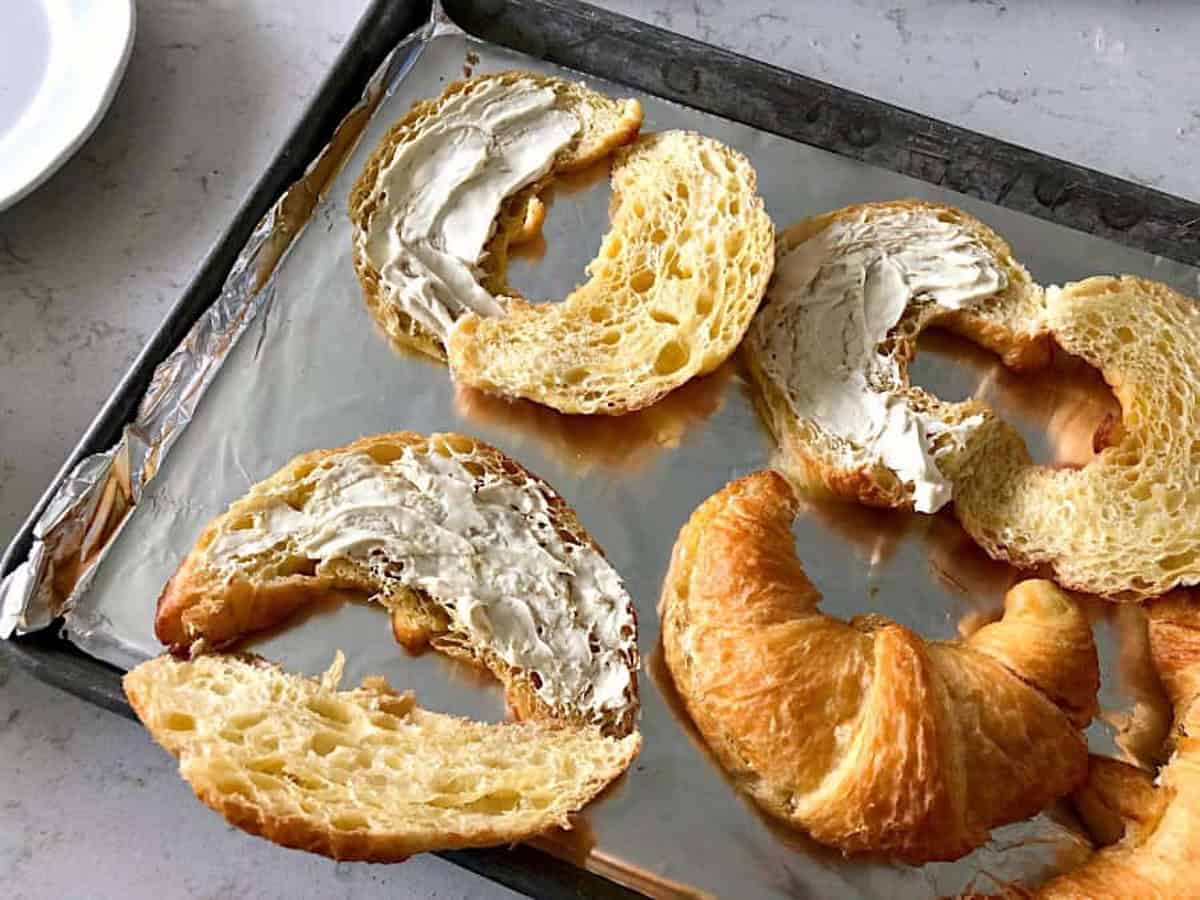 Sliced croissants with cream cheese spread on a foil-lined baking tray.