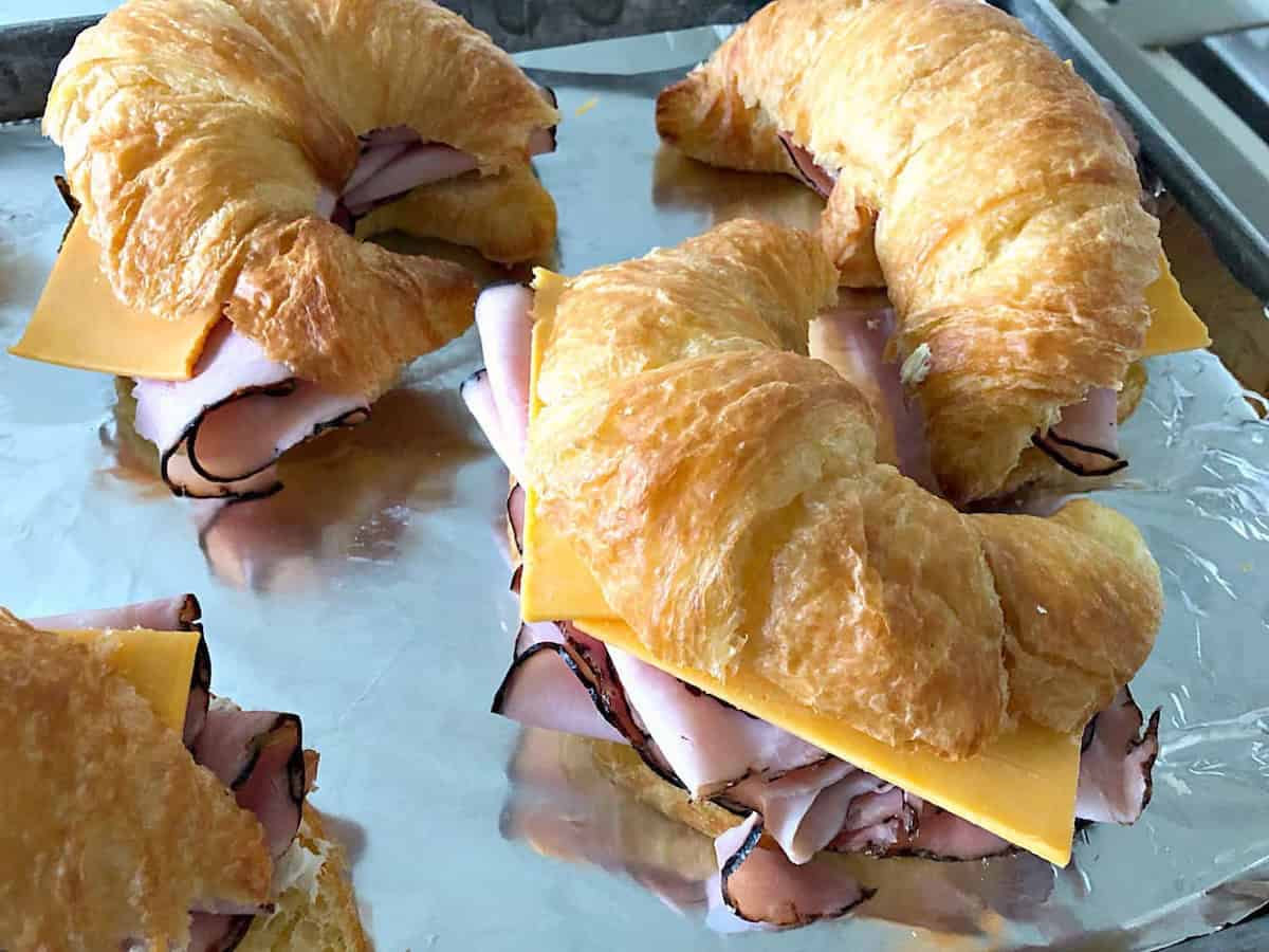 Four croissant sandwiches with ham and cheese on foil-lined baking sheet.