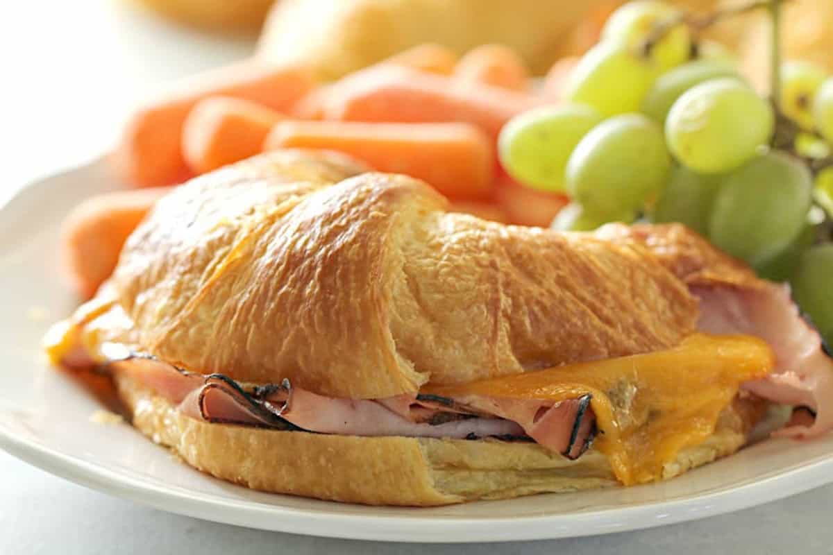 A croissant sandwich with ham and cheese is served on a white plate alongside green grapes and carrot sticks.