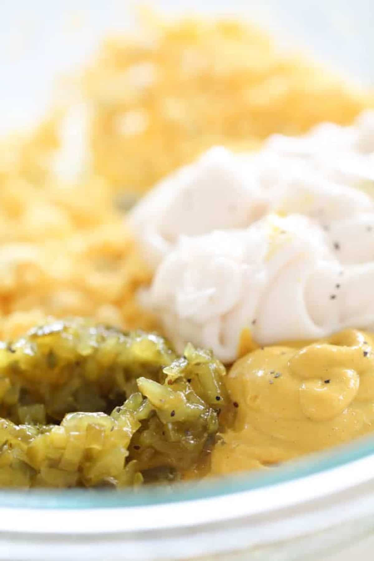 Close-up of a glass bowl containing chopped pickles, mustard, mayonnaise, and shredded cheese.