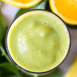 A glass of green smoothie seen from above, with fresh spinach leaves and halved oranges in the background.