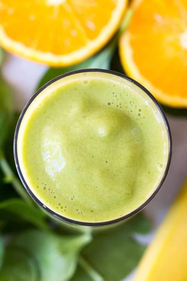 A glass of green smoothie seen from above, with fresh spinach leaves and halved oranges in the background.